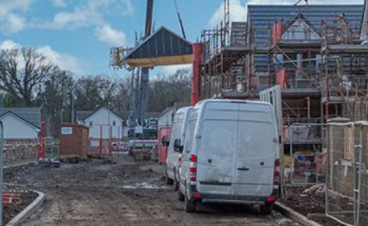 Modern Scottish Residential Building Site at various levels of construction and completion surrounded by scaffolding fencing and other materials. A Pre-constructed Roof is being lifted with a Crane.