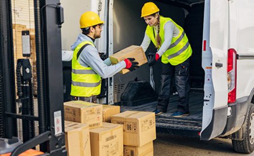 Two warehouse workers transfer cardboard boxes from a forklift to a delivery van A male worker in a reflective uniform passes cardboard boxes to a female colleague standing in a van and arranges them and prepares them for delivery