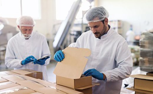 Young Caucasian employee in sterile uniform packing goods in boxes. In background supervisor holding tablet and counting boxes.